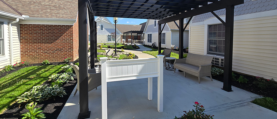 Ohio Living Cape May memory care at Agape Crossing featuring an outdoor courtyard with a black pergola, seating area, and raised planter box. Surrounding buildings display brick and light‑colored siding with gabled roofs. The landscaped walkway includes green lawns, hostas, and flowers, creating a peaceful, secure environment for residents living with dementia or Alzheimer’s.