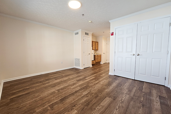 Ohio Living Cape May assisted living apartment featuring a bright, compact interior with wood‑grain flooring, light beige walls, and a kitchenette with oak cabinets, sink, microwave, and mini‑refrigerator. A large window at the far end fills the living area with natural light, creating a warm, comfortable environment for residents.