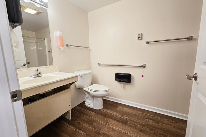 Ohio Living Cape May assisted living apartment bathroom featuring accessible design with beige walls, wood‑grain flooring, and a vanity sink with mirror. The space includes a toilet with stainless steel grab bars, wall‑mounted soap dispenser, and emergency pull cord, providing a safe and comfortable environment for senior residents.