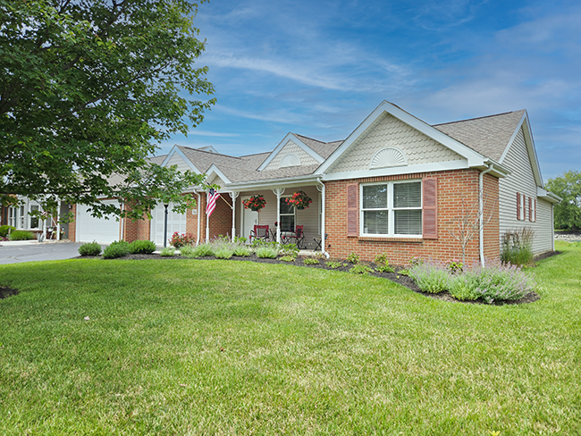 Single‑story brick and siding home with gabled roof, manicured lawn, and landscaped garden beds. The front porch features white columns, hanging flower baskets, seating, and an American flag, creating a welcoming suburban appearance under a bright blue sky.