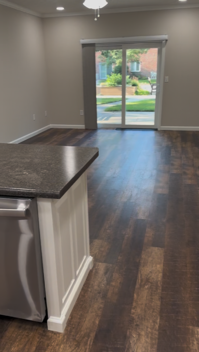 Modern apartment interior: Open living space with dark wood floors, neutral walls, and recessed lighting. A kitchen island with a dark countertop and stainless steel appliance sits in the foreground, while sliding glass doors lead to a sunny patio with greenery outside.