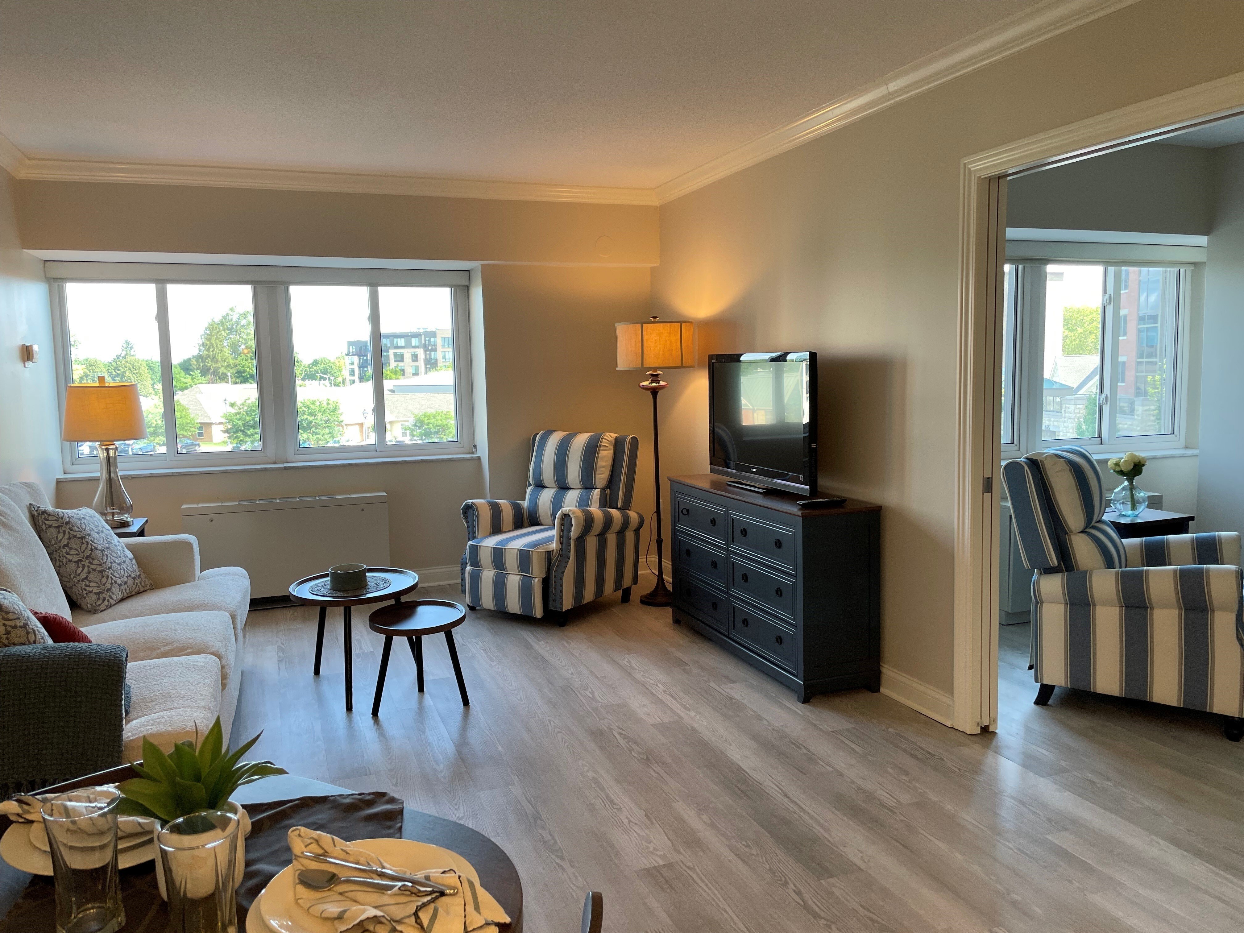 Bright living room interior: Spacious living area with large windows, striped armchairs, a white sofa, and a dark TV stand. Decor includes lamps, small tables, and neutral walls, creating a cozy, modern apartment atmosphere with natural light and wood‑style flooring.
