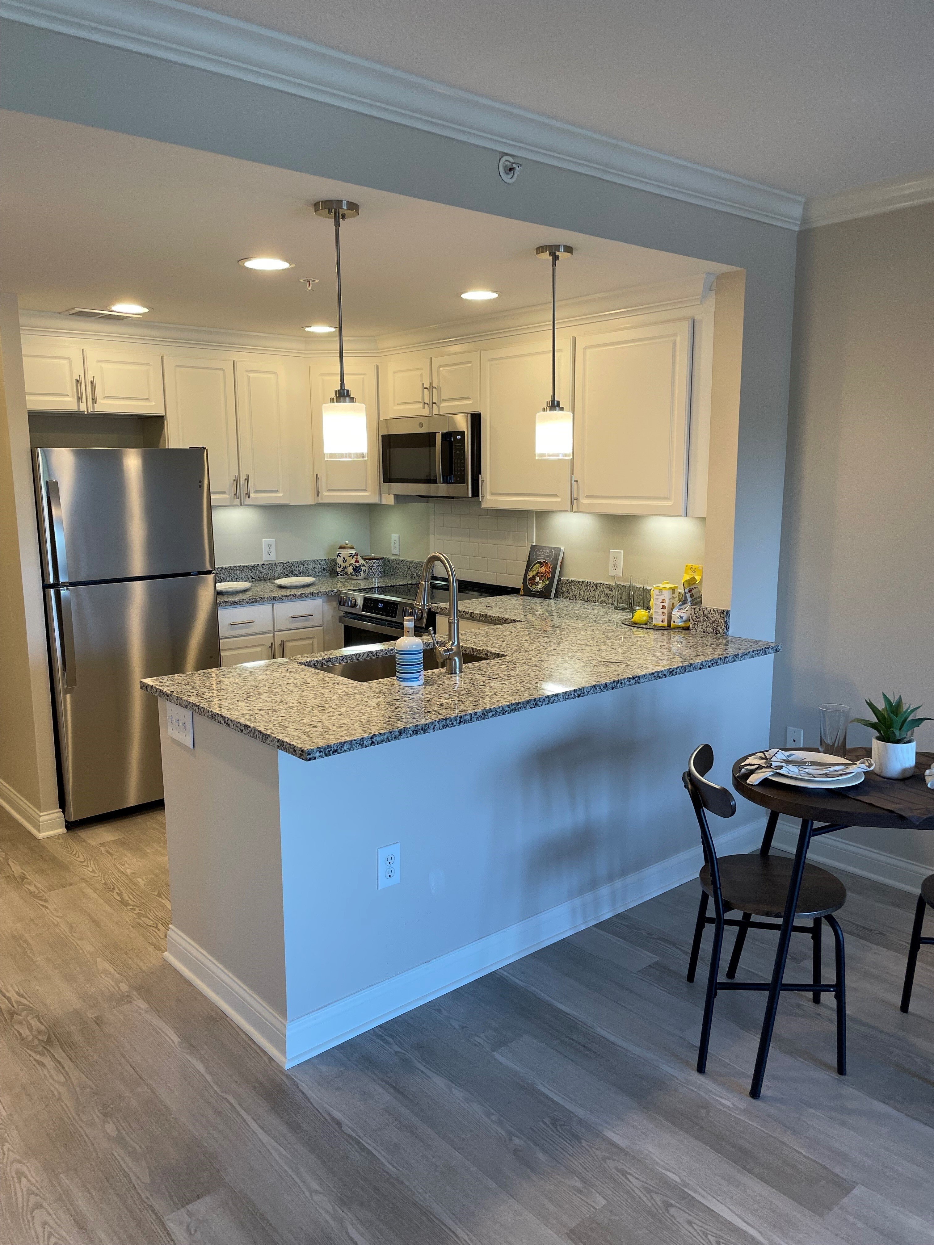 Modern kitchen interior: Bright kitchen with white cabinets, granite countertops, stainless steel appliances, pendant lighting, and a small dining table with two chairs, showcasing a clean, contemporary apartment design with neutral tones and wood‑style flooring.