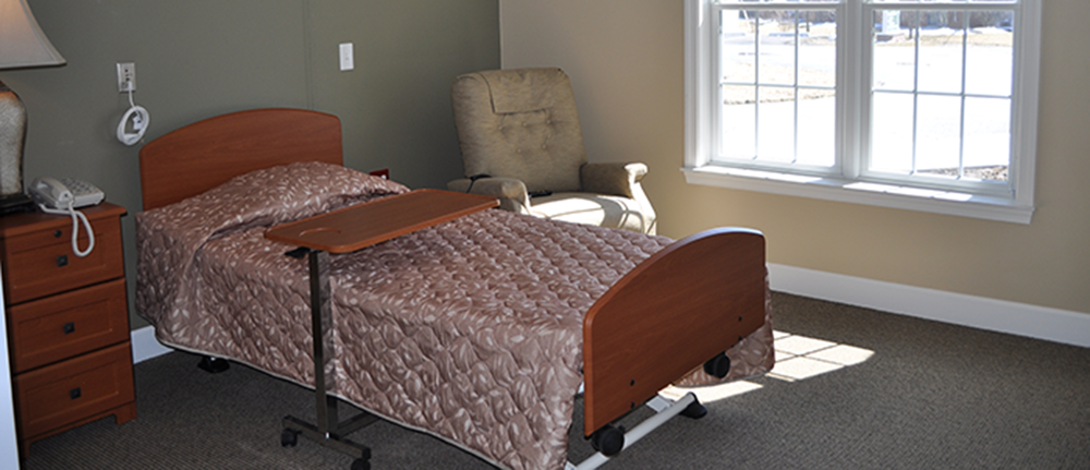 Ohio Living Cape May skilled short‑term rehab room featuring a single adjustable bed with wooden headboard and footboard, overbed table, and bedside nightstand with lamp and telephone. The space includes a comfortable armchair near a large window providing natural light, creating a clean, restful environment designed for recovery and rehabilitation.