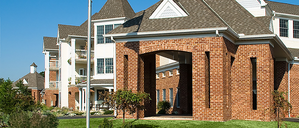 Ohio Living Cape May Cambridge Apartments featuring a multi‑story brick and white‑sided building with gabled roofs and large windows. The structure includes a covered brick entrance, balconies, and landscaped grounds with green lawns and shrubs under a clear blue sky.