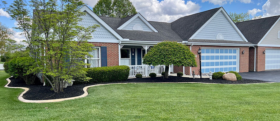 Ohio Living Cape May independent living villa featuring a single‑story brick and light blue‑gray exterior with white trim and dark gray gabled roof. The home includes a welcoming front porch with white railings, manicured lawn, curved mulch beds, and neatly trimmed shrubs and trees under a partly cloudy sky.