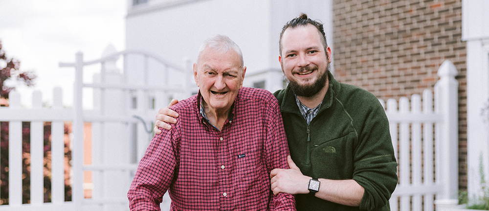 An older man and a younger man stand together outside at Agape Crossing, Ohio Living Westminster-Thurber, smiling warmly in front of a white picket fence and brick building. The older man wears a red plaid shirt, while the younger man in a green pullover has his arm around him, showing friendship and support. The image reflects Ohio Living’s commitment to compassionate care, intergenerational connection, and community engagement within its memory care program.