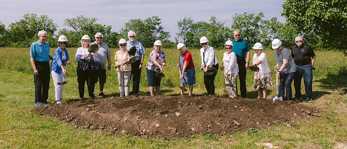 Group of senior adults and staff wearing white construction helmets participating in a ceremonial groundbreaking outdoors at Ohio Living Mount Pleasant. The group stands on a grassy field behind a patch of freshly turned soil under a sunny blue sky, symbolizing community growth and new development.